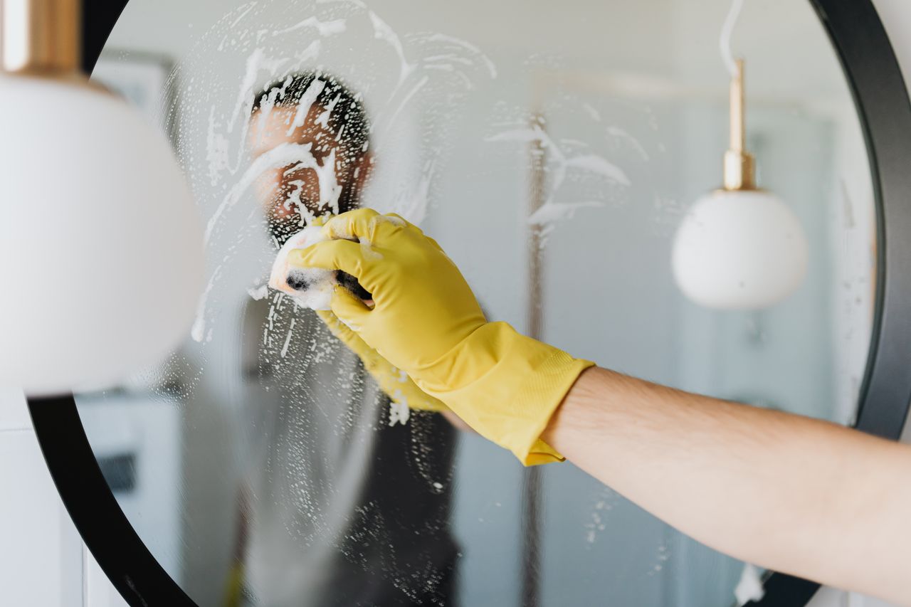 services-01 Anonymous male in yellow rubber gloves wiping foam from round mirror in bathroom